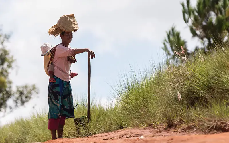 DEICHMANN-Stiftung | Madagascar Mobile Clinic – auf einen Arzt kommen in Madagaskar ca. 5.000 Einwohner – Frau mit Kleinkind auf Rücken gebunden und einer Last auf dem Kopf steht an der Straße Madagascar Mobile Clinic – Frau und Kind stehen am Straßenrand – von den wenigen Kilometern Straße, die Madagaskar überhaut hat, ist nur ein Bruchteil asphaltiert