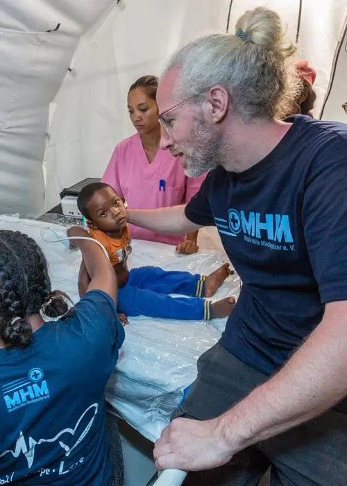 DEICHMANN-Stiftung | Madagascar Mobile Clinic – medizinisches Personal befähigen – Dr. Fabian Bendel unterstützt eine madagassische Ärztin, die sich mit dem Ultraschallgerät vertraut macht Madagascar Mobile Clinic – Ärztin macht Ultraschall bei Kind, Arzt hält Jungen fest und blickt mit Ärztin auf Bildschirm